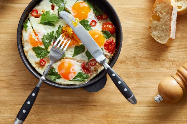 Top view of fried eggs with parsley and chili pepper on wooden table with cutlery and bread — Stock Photo