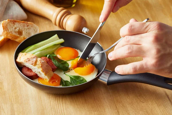 Vista recortada del hombre comiendo huevos fritos con tenedor y cuchillo en la mesa de madera - foto de stock