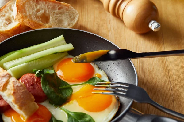 Spiegeleier in der Pfanne mit Gemüse und Wurst neben Gabel, Messer, Brot am Holztisch — Stockfoto