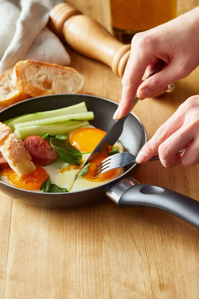 Vista recortada de la mujer comiendo huevos fritos con tenedor y cuchillo en la mesa de madera - foto de stock