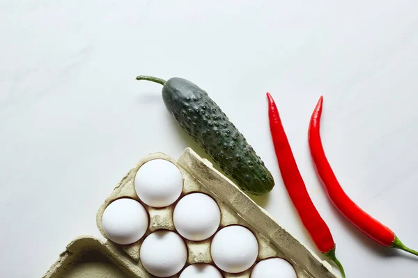 Top view of raw chicken eggs in box near cucumber and chili peppers on marble surface — Stock Photo