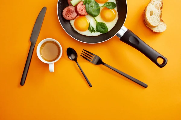 Top view of fried eggs with spinach leaves, cucumber and sausage in frying pan near coffee, cutlery and bread on orange background — Stock Photo