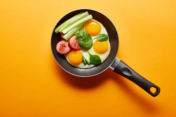 Top view of fried eggs with spinach leaves, cucumber and sausage in frying pan on orange background — Stock Photo