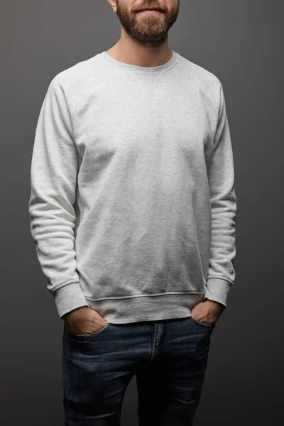 Cropped view of bearded man in blank basic grey sweatshirt with hands in pockets on black background — Stock Photo