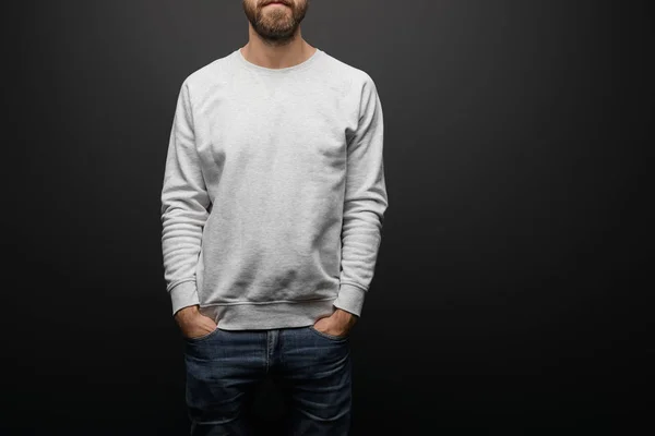 Partial view of bearded man in blank basic grey sweatshirt with hands in pockets isolated on black — Stock Photo