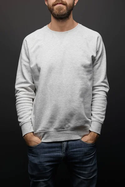 Partial view of bearded man in blank basic grey sweatshirt with hands in pockets isolated on black — Stock Photo