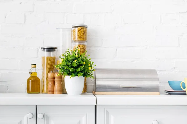 Intérieur moderne de cuisine blanche avec boîte à pain, pâtes, huile et plante verte près du mur de briques — Photo de stock