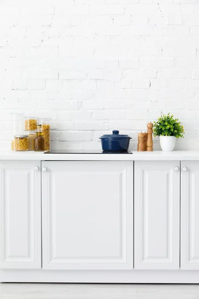 Modern white kitchen interior with pot on electric induction cooktop near decor and food containers near brick wall — Stock Photo