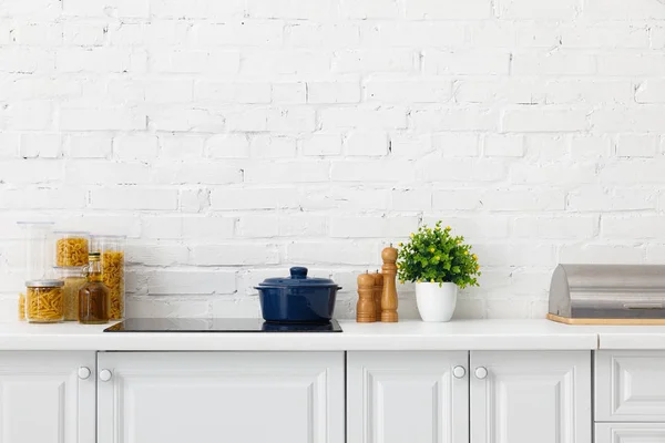 Intérieur moderne de cuisine blanche avec casserole sur table de cuisson à induction électrique près des conteneurs de plantes et d'aliments près du mur de briques — Photo de stock