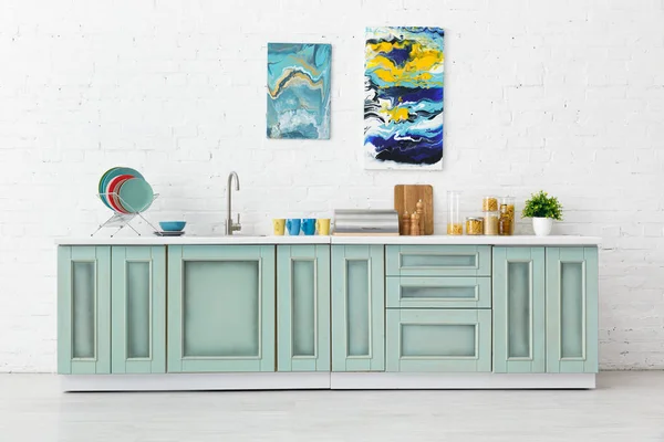 Современный белый и бирюзовый интерьер кухни с посудой и абстрактными картинами на кирпичной стене — стоковое фото