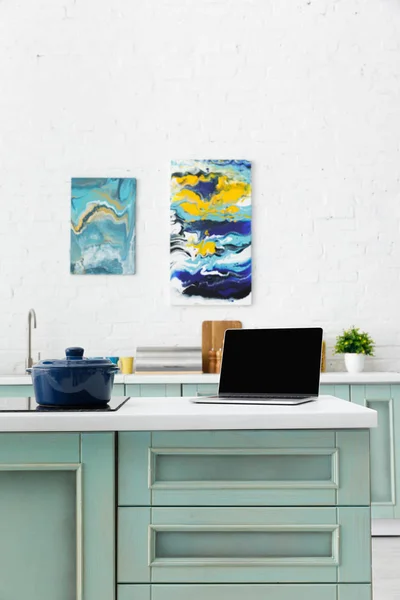 Selective focus of laptop and pot on induction cooktop with kitchenware and abstract paintings on background — Stock Photo