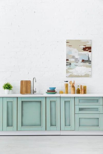Interior de la cocina blanca y turquesa con utensilios de cocina y pintura abstracta en la pared de ladrillo - foto de stock