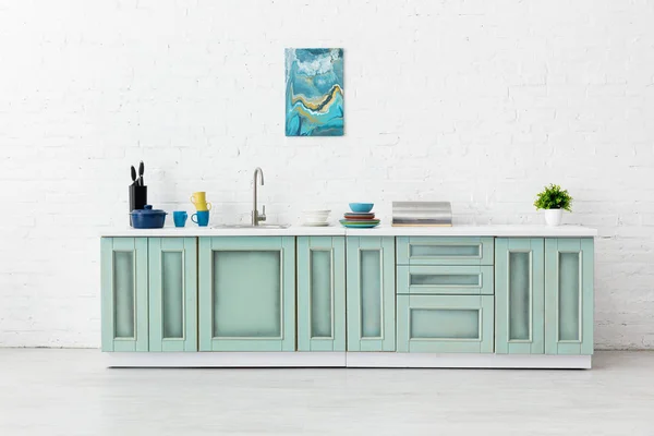 Cocina blanca y turquesa interior con fregadero, vajilla y pintura abstracta en la pared de ladrillo - foto de stock