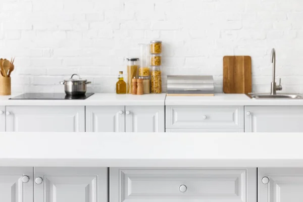 Enfoque selectivo de mesa vacía y minimalista moderno interior de cocina blanca con utensilios de cocina y alimentos cerca de la pared de ladrillo en el fondo - foto de stock