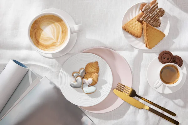 Vista superior de galletas con tazas de café y revistas sobre tela blanca - foto de stock