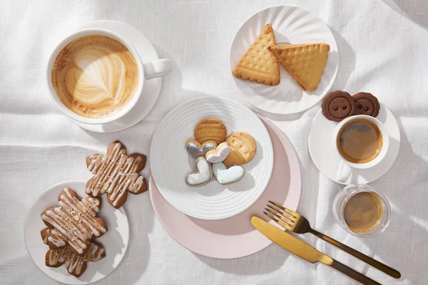 Draufsicht auf leckere Kekse mit Glasur und Kaffee auf weißer Tischdecke — Stockfoto