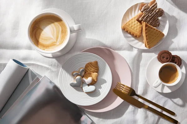 Vue du dessus des tasses à café à côté des biscuits et des magazines sur la nappe blanche — Photo de stock