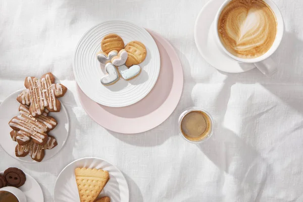 Draufsicht auf Kekse mit Glasur und Tassen Kaffee auf weißer Tischdecke — Stockfoto