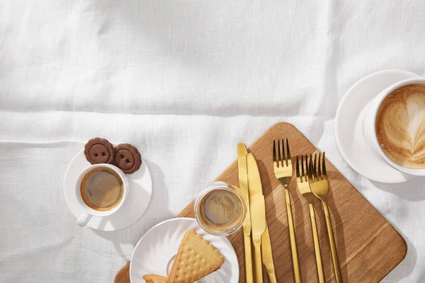 Vista superior de tazas y vaso de café con galletas y cubiertos sobre tabla de cortar en tela blanca - foto de stock