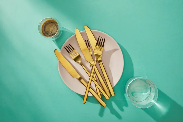 Vue de dessus des couverts dorés sur assiette avec café et eau dans des verres sur surface turquoise — Photo de stock