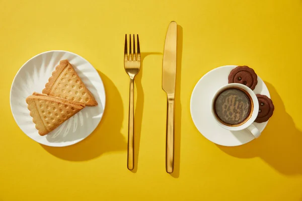 Blick von oben auf goldenes Besteck mit Kaffeetasse und Keksen auf gelbem Hintergrund — Stockfoto