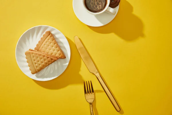 Draufsicht auf Tasse Kaffee mit goldenem Besteck und leckeren Keksen auf gelber Oberfläche — Stockfoto