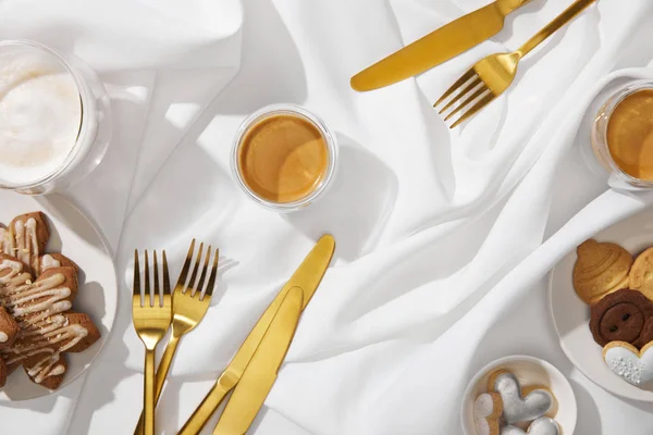 Blick von oben auf leckere Kekse mit frischem Kaffee und goldenem Besteck auf weißem Tuch — Stockfoto