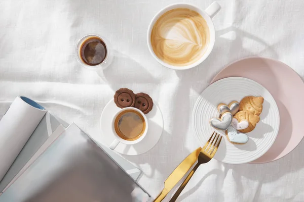 Vista superior de biscoitos saborosos com café fresco ao lado de revistas na toalha de mesa branca — Fotografia de Stock