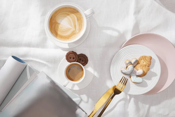 Draufsicht auf Gourmet-Kekse mit Tassen Kaffee und Zeitschriften auf weißer Tischdecke — Stockfoto