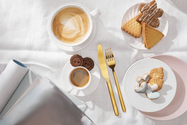 Vista superior de galletas recién horneadas con café y revistas sobre tela blanca - foto de stock