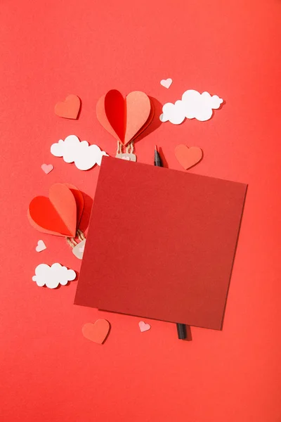 Top view of paper heart shaped air balloons in clouds near blank card and pencil on red background — Stock Photo