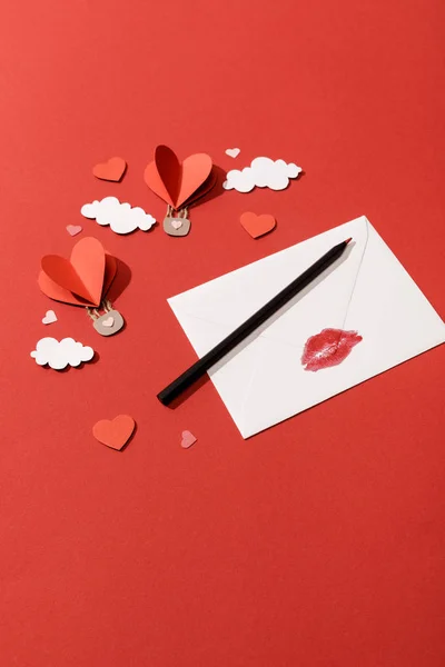 Paper clouds and heart shaped air balloons, envelope with lip print and pencil on red background — Stock Photo