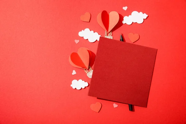 Top view of paper heart shaped air balloons in clouds near blank card and pencil on red background — Stock Photo