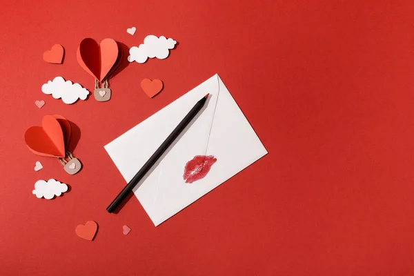 Top view of paper clouds and heart shaped air balloons, envelope with lip print and pencil on red background — Stock Photo
