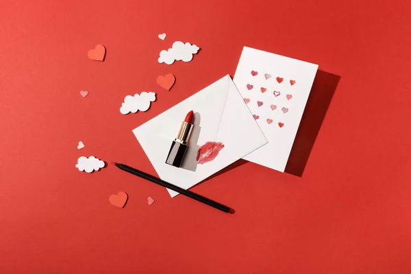 Vue du dessus des nuages de papier et des coeurs, carte de vœux près de l'enveloppe avec imprimé lèvre, rouge à lèvres et crayon sur fond rouge — Photo de stock