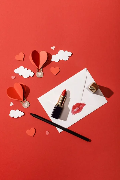 Вид зверху паперових хмар і повітряних кульок у формі серця, конверт з принтом для губ, помадою та олівцем на червоному тлі — стокове фото