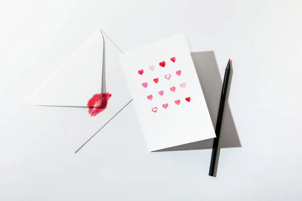 Вид поздравительной открытки с сердечками у конверта с отпечатком губы и карандашом на белом фоне — Stock Photo