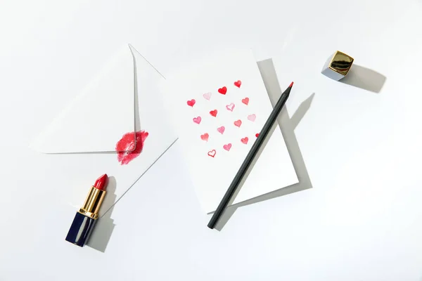 Top view of greeting card with hearts near envelope with lip print, lipstick and pencil on white background — Stock Photo