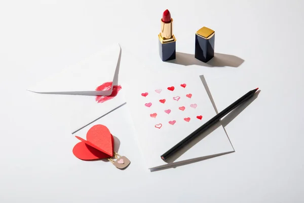 Tarjeta de felicitación con corazones cerca del sobre con impresión de labios, lápiz labial y lápiz sobre fondo blanco - foto de stock