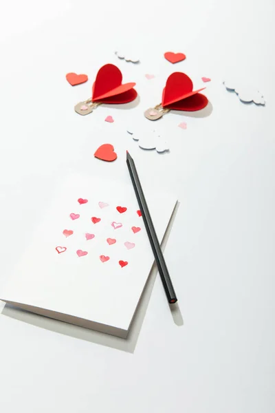 Selective focus of greeting card with hearts and pencil near paper heart shaped air balloons in clouds on white background — Stock Photo