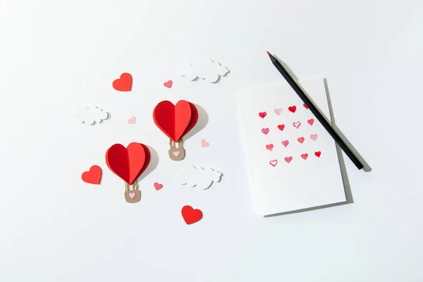 Top view of greeting card with hearts and pencil near paper heart shaped air balloons in clouds on white background — Stock Photo