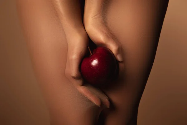 Cropped view of woman in nylon tights holding ripe red apple isolated on brown — Stock Photo