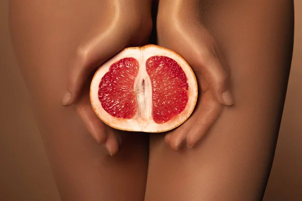 Abgeschnittene Ansicht einer Frau in Nylonstrumpfhosen, die Grapefruit halb isoliert auf braun hält — Stockfoto