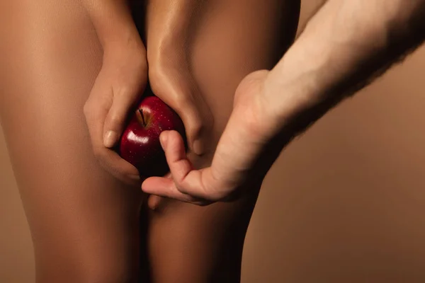 Vista recortada del hombre que llega a la mujer en medias de nylon sosteniendo manzana roja madura aislada en marrón - foto de stock