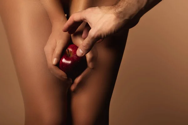 Vue recadrée de l'homme atteignant la femme en collants en nylon tenant pomme rouge mûre isolé sur brun — Photo de stock