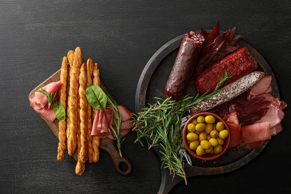 Vue de dessus de délicieux plateaux de viande servis avec des olives, des bâtonnets de pain et des herbes sur des planches sur une table noire en bois — Photo de stock