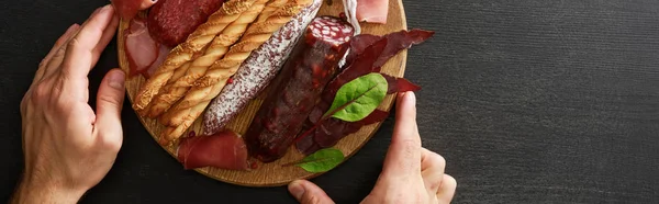 Обрезанный вид человека с вкусным мясным блюдом подается с хлебными палочками и травами на борту на деревянном черном столе, панорамный снимок — стоковое фото