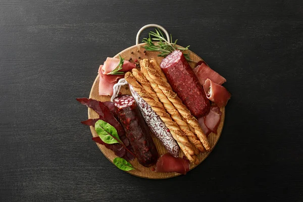 Top view of delicious meat platter served with breadsticks and herbs on board on wooden black table — Stock Photo