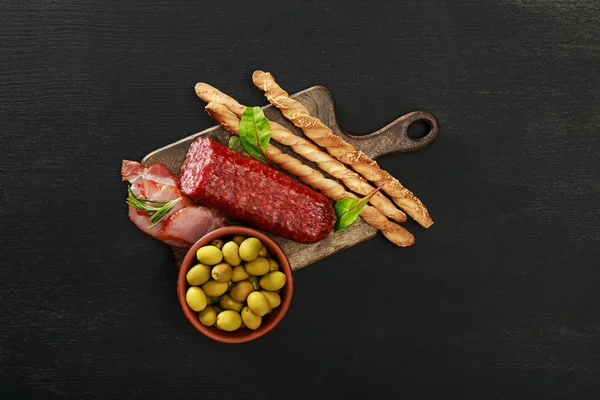 Vue de dessus du délicieux plateau de viande servi avec des olives et des grissini à bord sur une surface noire — Photo de stock
