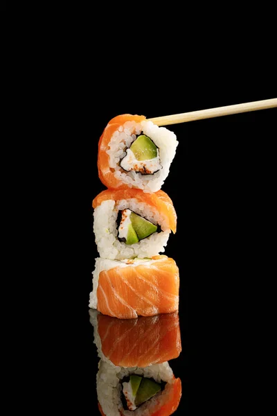 Chopstick perto de delicioso sushi Filadélfia com abacate, queijo cremoso, salmão e masago caviar isolado em preto com reflexão — Fotografia de Stock
